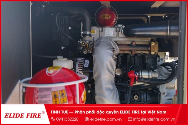 Ảnh chụp thật: cận cảnh bóng chữa cháy Elide Fire TECHIDEAS lắp cố định trên khung kim loại phía trên tủ điều khiển máy phát điện diesel trong nhà xưởng, thấy rõ mũi tên vàng hướng lên và lớp vỏ bóng màu đỏ, phía sau là dàn máy phát công nghiệp cỡ lớn phủ bụi công nghiệp nhẹ.